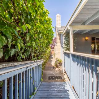 A narrow wooden walkway with blue railings, lined by lush greenery and leading to a house under a clear sky.