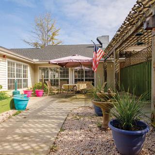 A cozy backyard patio featuring colorful potted plants, outdoor seating under a pergola, and a flag waving in the breeze on a sunny day.
