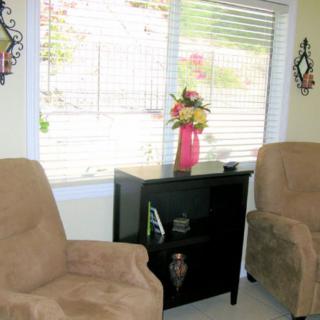 A cozy living room with two armchairs, a cabinet with a flower arrangement in a vase, and natural light streaming through the window blinds.