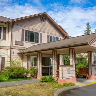A two-story residential building with a sheltered entrance, surrounded by greenery under a partly cloudy sky.