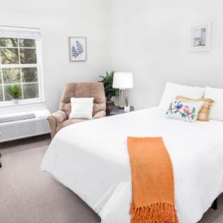 A bright and cozy bedroom featuring white bedding, a comfortable armchair, and natural light streaming in from the window.