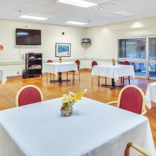A clean, bright dining room with neatly arranged tables and chairs, featuring a television on the wall and floral centerpieces.