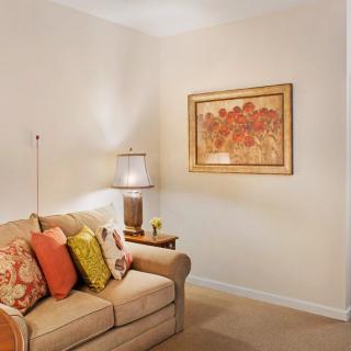 A cozy living room corner featuring a beige sofa with colorful pillows, a lamp on a side table, and framed artwork on the wall.