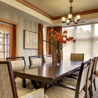 A spacious dining room featuring a large table surrounded by chairs, with elegant lighting from a chandelier and bright natural light streaming in through the windows.