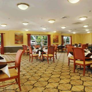 A well-lit dining room with arranged tables and chairs, featuring large windows and carpeted flooring.
