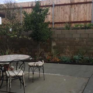 A backyard patio with a table and chairs, surrounded by a garden and wooden fence.