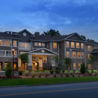 A well-lit residential building in the evening with landscaped surroundings and a road in the foreground.