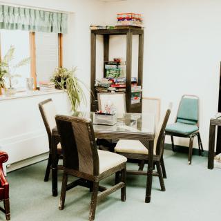 A cozy dining room with a wooden table and chairs, plants on the windowsill, and a tall shelf in the corner.