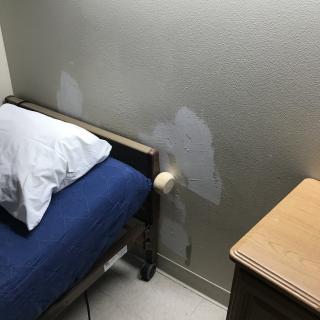 A bedroom with a bed, pillow, and patched wall beside a side table.