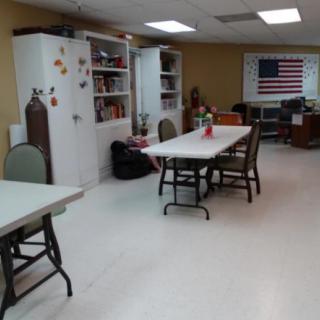 A classroom with tables, chairs, and shelves, featuring an American flag on the wall.