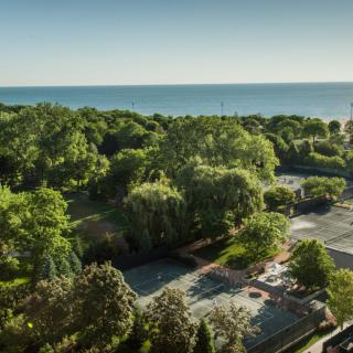 A scenic view of tennis courts nestled among lush green trees with a lake in the background.