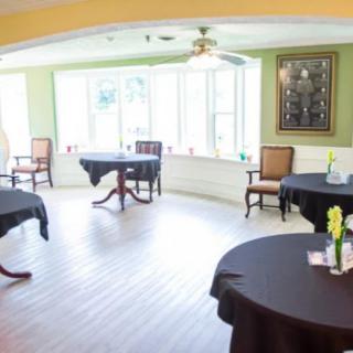 A bright room with round tables covered in black tablecloths and chairs around them, illuminated by natural light from large windows.