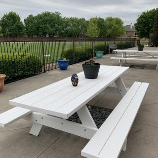 A white picnic table and benches on a patio, adorned with potted plants, overlooking a green field.