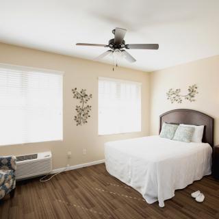 A bright and minimalist bedroom with a neatly made bed, ceiling fan, and decorative wall art.