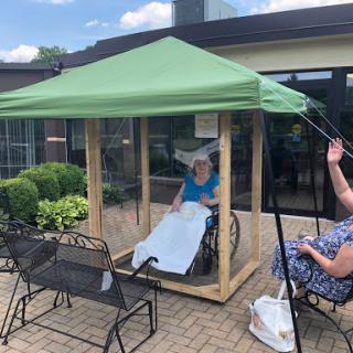 An elderly person in a wheelchair enjoying some outdoor time under a canopy, while a caregiver attends to them.