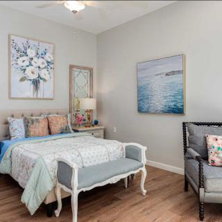 A cozy bedroom with elegant decor, featuring a bed with pastel pillows, artwork on the walls, and a comfortable chair.