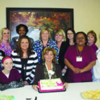 A group of people gathered around a table with a cake, celebrating in an office setting.