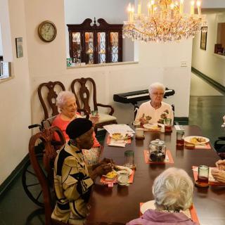 A group of elderly people sitting around a table enjoying a meal together in a dining room with a chandelier.