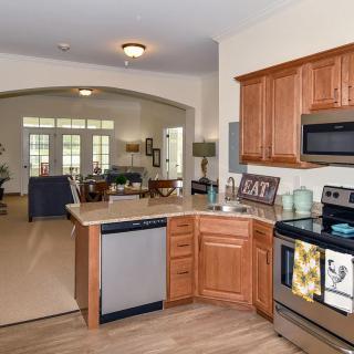 A modern kitchen with wooden cabinets and stainless steel appliances, featuring an open layout leading to the dining and living areas.