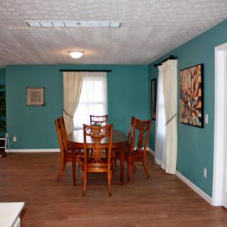 A cozy dining room with teal walls features a wooden table surrounded by chairs, hardwood flooring, and adorned with artwork and curtains.