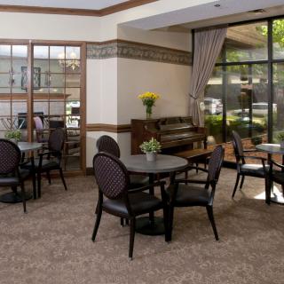A cozy lounge area with tables, chairs, and a piano, featuring large windows and elegant curtains.