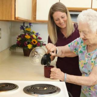 An elderly woman pouring coffee with the help of a caregiver in a kitchen setting.