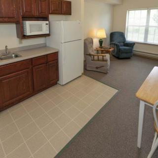 A cozy apartment interior featuring a kitchen with cabinets, microwave, and refrigerator alongside a living area with armchairs and a window.