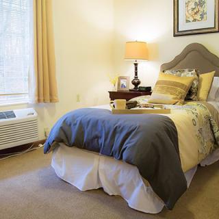 A cozy bedroom with a neatly made bed, warm lighting from a lamp, and a large window letting in natural light.