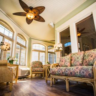 A cozy sunroom featuring rattan furniture with floral upholstery, illuminated by natural light through large windows and a decorative ceiling fan.