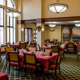 An elegant dining area with multiple tables and chairs, illuminated by large windows and chandeliers.