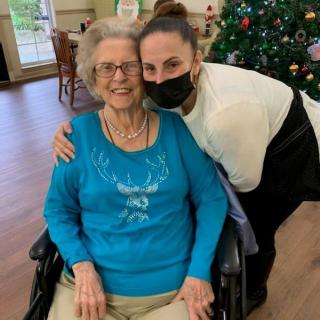 An elderly woman sitting in a wheelchair is hugged by a younger woman wearing a mask, with a Christmas tree in the background.