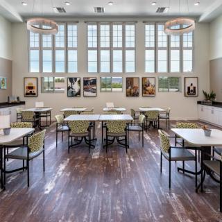 A modern dining room with large windows, tables, and chairs arranged neatly, featuring artwork on the walls and wooden flooring.