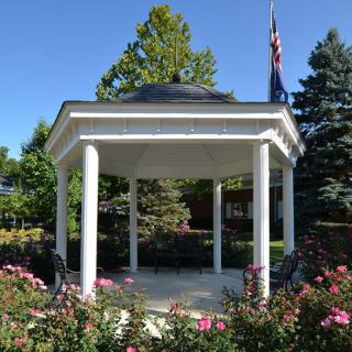A white gazebo surrounded by a garden with blooming flowers and lush trees.
