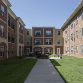 A three-story apartment building with a brick facade, featuring a central pathway and surrounding lawn.