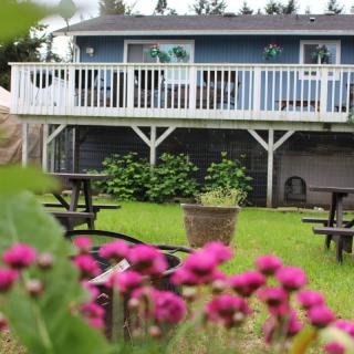 A cozy house with a wooden deck overlooks a garden with vibrant pink flowers and picnic tables on the lawn.