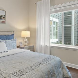 A cozy bedroom with a neatly made bed, a bedside lamp, and curtains framing a large window.
