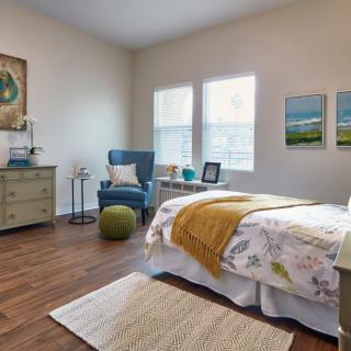 A cozy and well-decorated bedroom with a bed, armchair, dresser, and two windows.