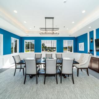 A modern dining room with blue walls and a large table surrounded by chairs, illuminated by a central chandelier.