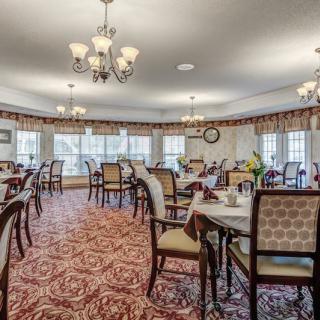An elegant dining room with multiple tables and chairs, illuminated by chandeliers, featuring carpeted flooring and large windows.