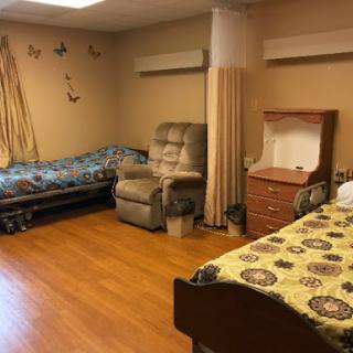 A spacious and comfortable shared room in a senior living facility, featuring beds, chairs, and warm decor.