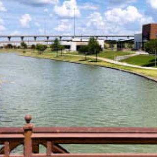 A scenic view of a park with a lake, bridge, and buildings under a partly cloudy sky.
