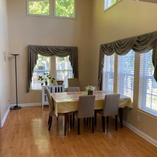 A bright dining room with a table set for six, featuring large windows with decorative curtains and wood flooring.
