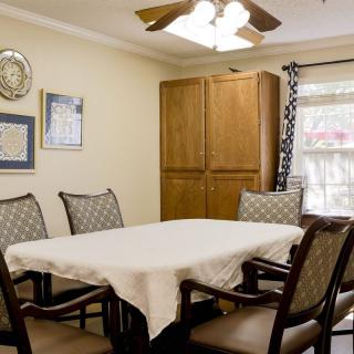 A cozy dining room with a table covered by a white cloth and surrounded by wooden chairs, featuring wall artwork and a ceiling fan overhead.