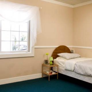 A simple bedroom featuring a single bed next to a window with a nightstand and lamp.