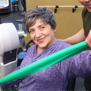 An elderly woman smiling while exercising with a resistance band, assisted by a therapist.