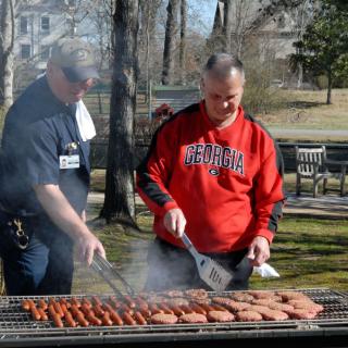 Two men grilling hot dogs and hamburgers outdoors in a park.