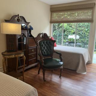 A cozy bedroom featuring a large armoire, an elegant green chair, a bedside lamp, and a window with garden view.