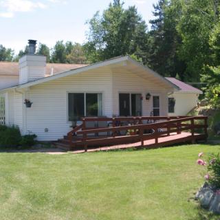 A single-story house with a wooden ramp leading to the patio, surrounded by a grassy yard and trees.