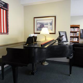 A grand piano placed in a room with wall artwork, a lamp, and a bookshelf.