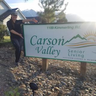 A person poses beside the Carson Valley Senior Living sign against a scenic backdrop.